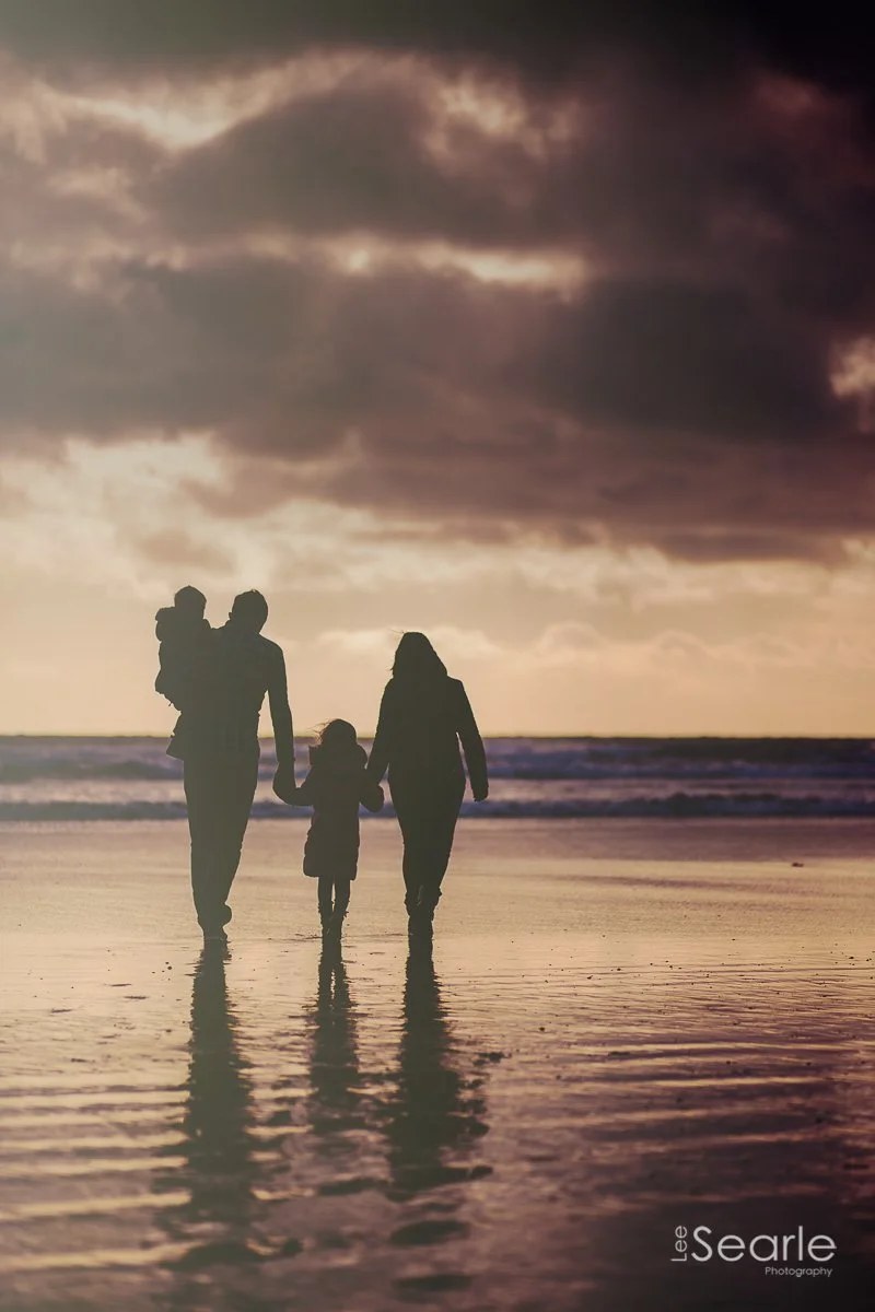 Family silhouetted walking along a Cornish beach at sunset, reflected in the wet sand