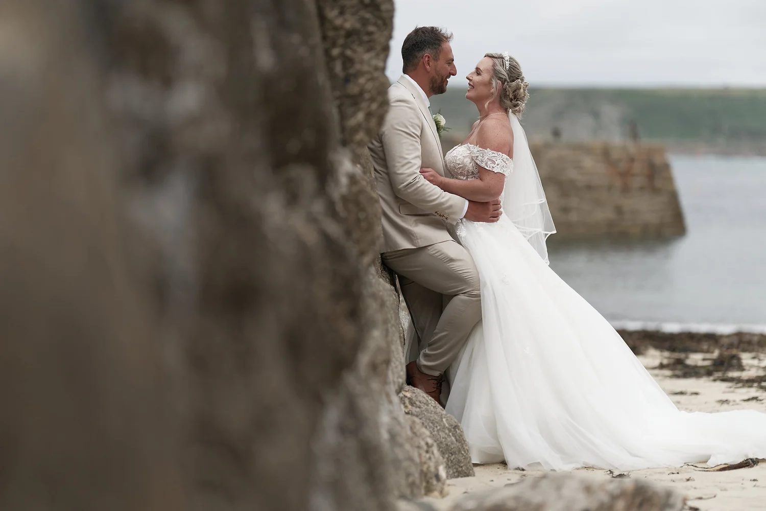 Bride and groom laughing against a granite wall on a Cornish beach