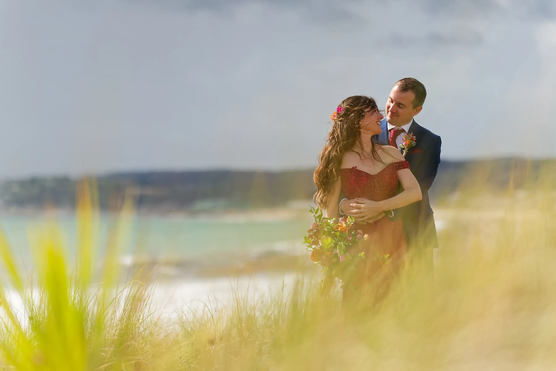 Bride and groom embracing among coastal grasses near Penzance after their wedding