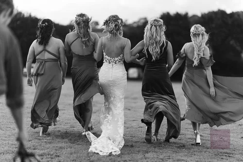 Bridesmaids walking arm in arm with the bride across a field, black and white